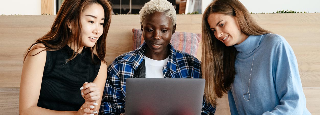 Three women sitting on a couch looking at a laptop.
